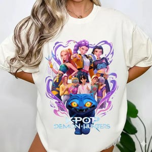 May include: Off-white t-shirt with a colourful graphic of animated characters, a blue cat, and the text "KPOP DEMON HUNTERS" in blue. The design incorporates purple and yellow elements.