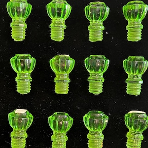 May include: A collection of twenty-one green glass light bulb-shaped objects with a threaded base. The objects are arranged in rows on a black surface. Some have a small white piece on top.