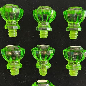 May include: A collection of eleven green glass cabinet knobs. Each knob has a faceted, rounded top and a threaded base for attachment. The knobs are arranged on a black surface, showcasing their bright, translucent color.