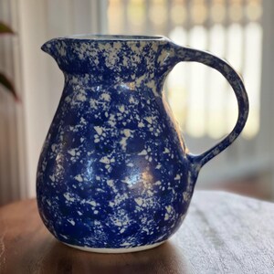 May include: A ceramic pitcher featuring a vibrant blue and white speckled pattern. The vessel has a rounded body, a curved handle on the right side, and a small pouring spout at the top rim, showcasing a consistent mottled finish throughout.