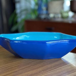 May include: A vibrant, glossy, blue ceramic bowl with a contrasting light blue interior. The bowl has a unique, angular shape and sits on a wooden surface. This decorative bowl is suitable for serving or display.