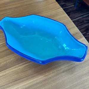 May include: A vibrant, turquoise-blue glass dish with a unique, elongated shape. The dish has a darker blue rim, creating a contrasting border. The glossy surface reflects light, highlighting its smooth texture. This decorative piece is suitable for serving or display.