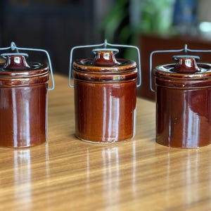May include: Three brown ceramic jars with lids and wire handles. The jars are cylindrical with a glossy finish. The lids have a small knob on top. The jars are arranged on a wooden surface.
