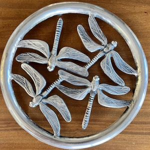 May include: A silver-colored, circular decorative piece featuring four dragonfly designs. The dragonflies are arranged within a ring, with detailed wings and bodies. The piece is likely made of metal and sits on a wooden surface.