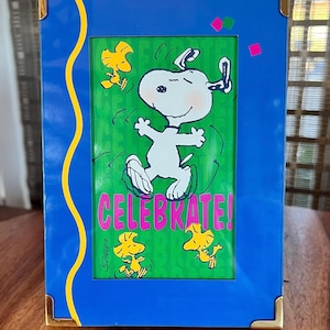 May include: A blue picture frame with gold corner accents and a yellow wavy design. The frame features a green background with Snoopy dancing and the word "CELEBRATE!" in pink. Yellow birds are also featured.