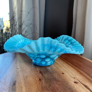 May include: A vintage, light blue glass bowl with a ruffled rim. The bowl features a hobnail pattern on the exterior and a scalloped edge. The bowl is displayed on a wooden surface, showcasing its elegant design.