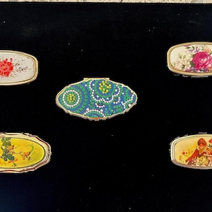 May include: A collection of five vintage pill boxes with ornate designs. The boxes feature floral patterns, geometric shapes, and a whimsical illustration. Each box has a gold-colored trim and a hinged lid, set against a black background.