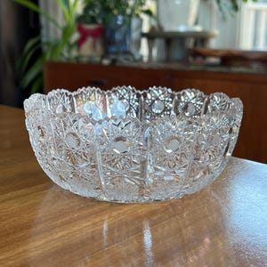 May include: A clear, cut-glass bowl with a scalloped rim. The bowl features intricate starburst and geometric patterns. The bowl is sitting on a wooden surface. This decorative bowl is ideal for serving or display.