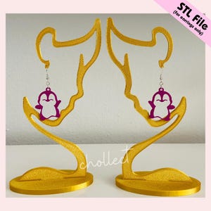 May include: Two gold-coloured earring display stands, each shaped like a stylised face, holding a pair of purple penguin earrings. The stands have a textured finish. The text "STL File (for earrings only)" is in the upper right corner.