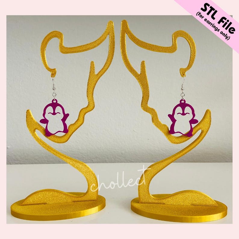 May include: Two gold-coloured earring display stands, each shaped like a stylised face, holding a pair of purple penguin earrings. The stands have a textured finish. The text "STL File (for earrings only)" is in the upper right corner.