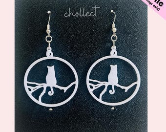 May include: White cat-shaped earrings. Each earring features a cat silhouette perched on a branch within a circular frame. The earrings have a silver hook and are displayed against a dark background. The text "chollect" is visible.
