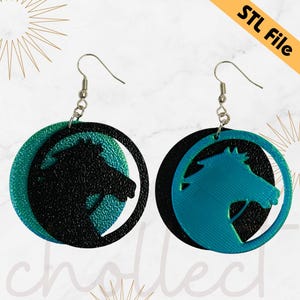 May include: A pair of black and teal blue earrings with a horse silhouette design. The earrings are made of a layered material with a textured finish. The earrings are attached to silver hooks.