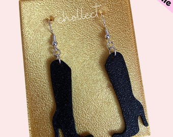 May include: A pair of black boot-shaped earrings with silver hooks. The earrings are displayed on a gold background.