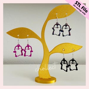May include: A gold-coloured earring display stand in the shape of a stylised tree, holding several pairs of penguin-shaped earrings. The earrings are in various colours, including purple, black, and a mix of blue and red. Text on the image reads "STL File (for earrings only)."