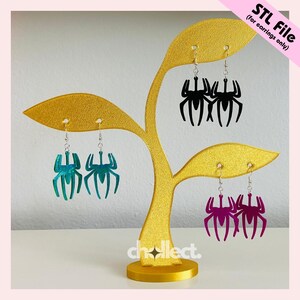 May include: A gold-colored earring display stand holding three pairs of spider-shaped earrings. The earrings are in teal, black, and purple. The text "STL File (for earrings only)" is in the upper right corner. The word "chellect" is at the bottom.