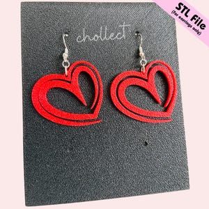 Puede incluir: Un par de pendientes rojos en forma de coraz&oacute;n con ganchos plateados. Los pendientes tienen un dise&ntilde;o en capas y est&aacute;n hechos de un material s&oacute;lido. Se muestran sobre una superficie negra texturizada. El texto "STL File (for earrings only)" est&aacute; en la esquina superior derecha.