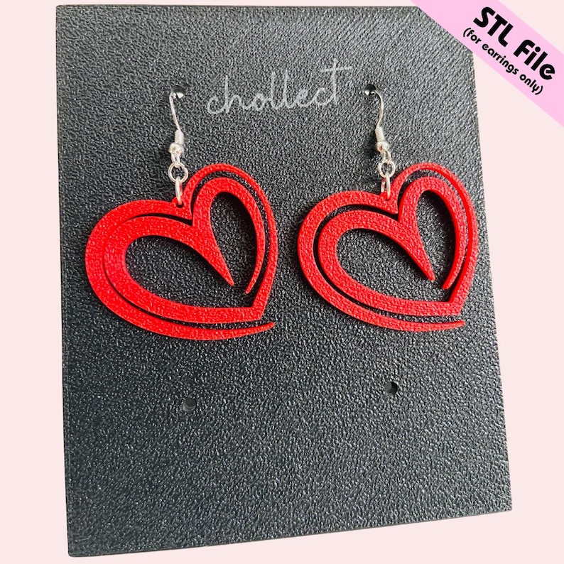 Puede incluir: Un par de pendientes rojos en forma de coraz&oacute;n con ganchos plateados. Los pendientes tienen un dise&ntilde;o en capas y est&aacute;n hechos de un material s&oacute;lido. Se muestran sobre una superficie negra texturizada. El texto "STL File (for earrings only)" est&aacute; en la esquina superior derecha.