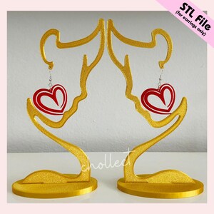 Puede incluir: Dos soportes dorados para pendientes, cada uno con la forma de una figura estilizada, que sostienen un pendiente rojo en forma de coraz&oacute;n. Los pendientes tienen un contorno blanco. El texto en la imagen dice "STL File (for earrings only)".