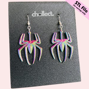 May include: A pair of iridescent spider earrings with silver hooks. The earrings are displayed on a textured black surface. The earrings have a rainbow color scheme, with hues of pink, blue, green, and yellow.