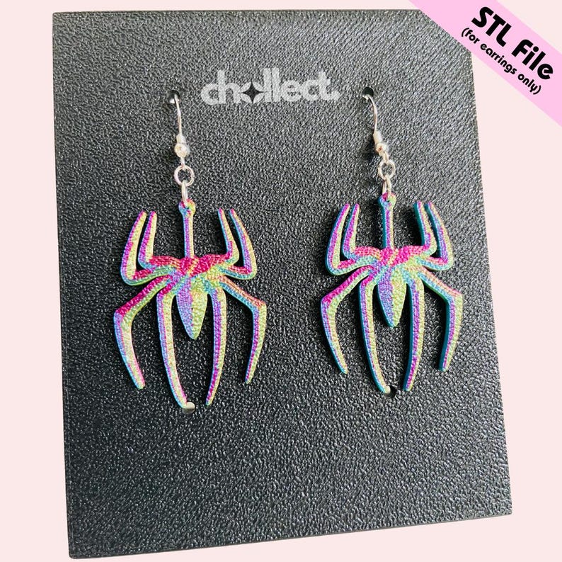 May include: A pair of iridescent spider earrings with silver hooks. The earrings are displayed on a textured black surface. The earrings have a rainbow color scheme, with hues of pink, blue, green, and yellow.