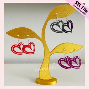 Puede incluir: Un expositor de pendientes dorado con tres pares de pendientes en forma de coraz&oacute;n. Los pendientes son rojos, iridiscentes y morados. El expositor tiene un dise&ntilde;o en forma de hoja. El texto en la imagen dice "STL File (for earrings only)" y "chollect".