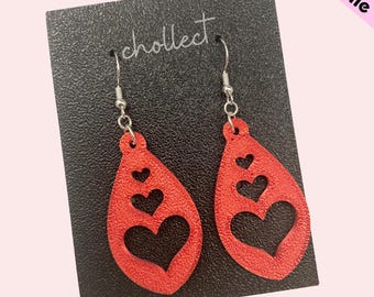 May include: A pair of red teardrop-shaped earrings with a heart cut-out design. The earrings are on a black background with the word "cholleect" written on it.
