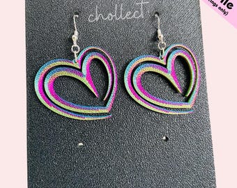 May include: Heart-shaped earrings with a rainbow iridescent finish. The earrings are layered with multiple colors, including pink, green, and blue. The earrings are hanging on a black display card. The text "chollect" is visible.