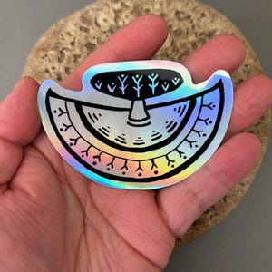 May include: A holographic sticker with a black and white design. The sticker features a stylized tree and a semi-circular shape with decorative elements. The sticker is held in a hand.
