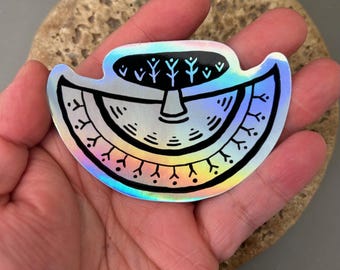 May include: A holographic sticker with a black and white design. The sticker features a stylized tree and a semi-circular shape with decorative elements. The sticker is held in a hand.