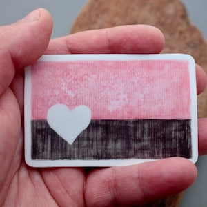 May include: A sticker with a pink and black design. The sticker features a white heart in the center of the pink section. The black section has a textured look.