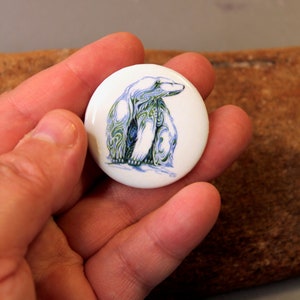 Op de afbeelding: Een witte knoop met een groene en blauwe illustratie van een ijsbeer. De beer zit en heeft een gedetailleerd ontwerp.