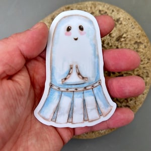 May include: A white ghost sticker with a blue and white skirt and a happy face. The ghost has pink cheeks and black eyes.