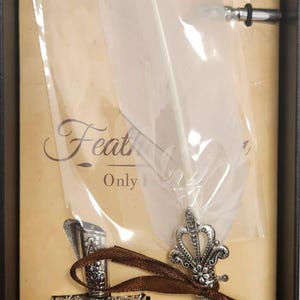 May include: A vintage-style calligraphy set in a black box. It includes a silver-colored quill pen with a crown design, a bottle of black ink, and several nibs. The set also features a decorative ring.