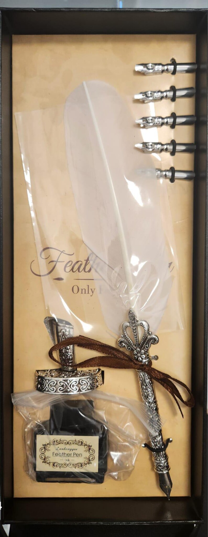 May include: A vintage-style calligraphy set in a black box. It includes a silver-colored quill pen with a crown design, a bottle of black ink, and several nibs. The set also features a decorative ring.