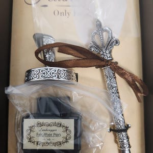 May include: A vintage-style calligraphy set featuring a silver-colored pen with ornate detailing, a matching inkwell, and a bottle of ink. The ink bottle has a label that reads "Lankongyue Feather Pen". The set is packaged in a box.