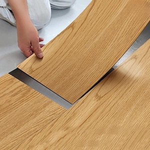 Op de afbeelding: Installatie van lichtbruine vloerbedekking met houtlook. Een hand plaatst een plank op een donkergrijze onderlaag. De planken hebben een houtnerfpatroon en zijn rechthoekig. De vloerbedekking wordt binnenshuis geïnstalleerd.