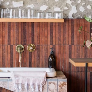 Puede incluir: Interior de baño con bañera blanca, grifos de latón y dispensador de jabón de cristal oscuro. La pared está alicatada con baldosas verticales rojas y marrones. Vasos transparentes sobre una estantería de madera. Una toalla rosa cuelga de la bañera.