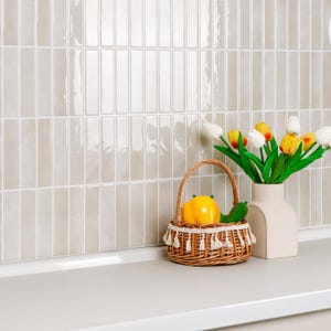Puede incluir: Una cesta de mimbre tejida con un borde de flecos decorativos que contiene un pimiento amarillo y un pepino verde, junto a un jarrón de cerámica beige lleno de tulipanes blancos, amarillos y naranjas frente a una pared de azulejos beige brillante.