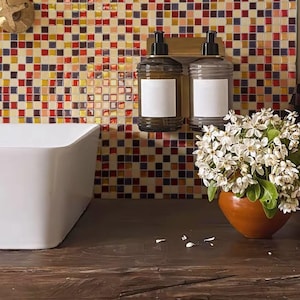 Puede incluir: Dos botellas de vidrio transparente con bombas, llenas de jabón líquido, montadas en un soporte de metal dorado contra una pared de mosaico de azulejos coloridos. Las botellas tienen etiquetas blancas sin texto.