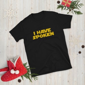 May include: Black short-sleeve t-shirt with the text "I HAVE SPOKEN" in yellow, a reference to a popular science fiction series. The shirt is displayed on a wooden surface with holiday decorations, including a red gift box and snowflake ornaments.