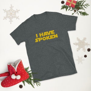 May include: Dark gray t-shirt with the text "I HAVE SPOKEN" in yellow. The shirt is surrounded by holiday decorations, including a red gift box, pine branches, and snowflake ornaments.