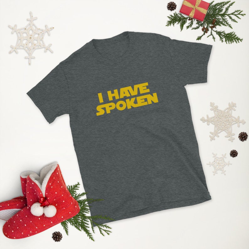 May include: Dark gray t-shirt with the text "I HAVE SPOKEN" in yellow. The shirt is surrounded by holiday decorations, including a red gift box, pine branches, and snowflake ornaments.