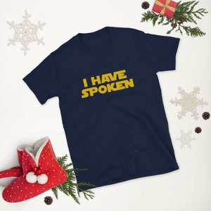 May include: Navy blue short-sleeve t-shirt with the text "I HAVE SPOKEN" in yellow, a design inspired by a popular science fiction franchise. The shirt is surrounded by holiday decorations, including a red gift box and snowflake ornaments.