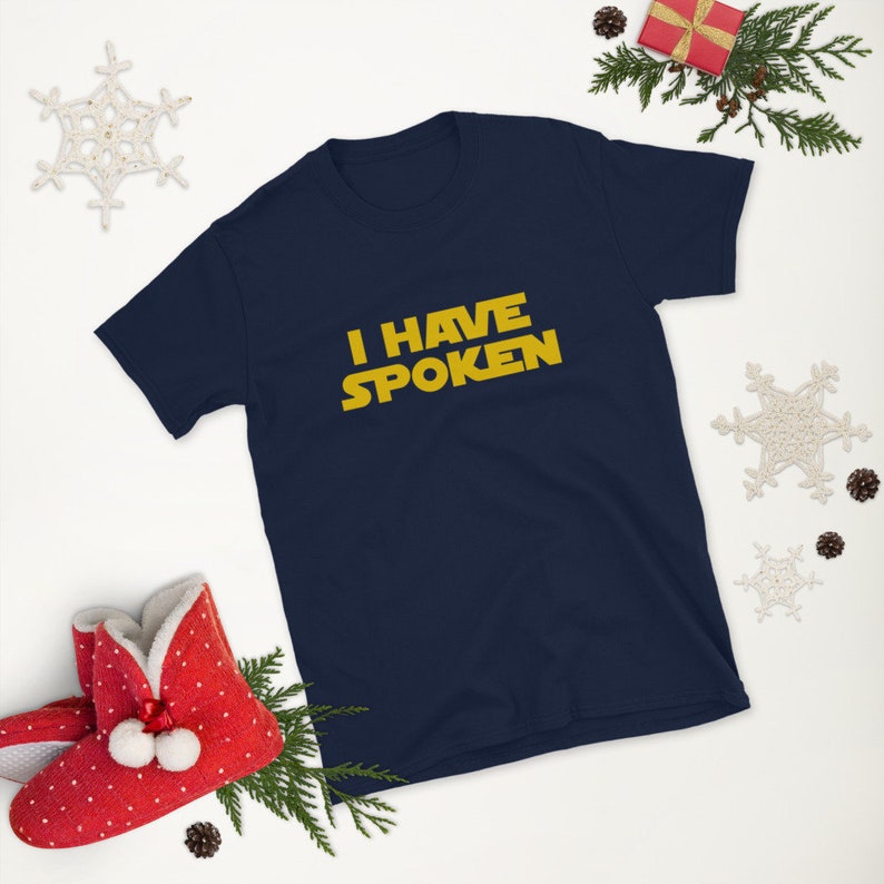 May include: Navy blue short-sleeve t-shirt with the text "I HAVE SPOKEN" in yellow, a design inspired by a popular science fiction franchise. The shirt is surrounded by holiday decorations, including a red gift box and snowflake ornaments.