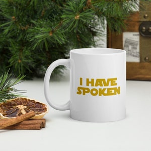 Puede incluir: Taza de cer&aacute;mica blanca con el texto dorado "I HAVE SPOKEN". La taza tiene un asa curva. El fondo incluye vegetaci&oacute;n y un cofre de madera.