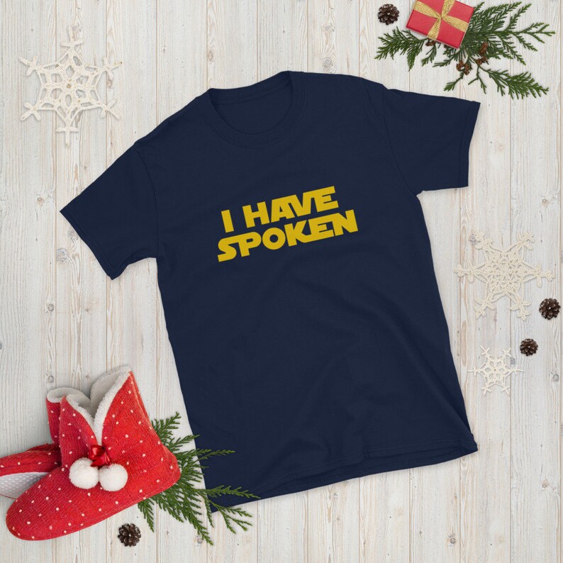 May include: Navy blue short-sleeve t-shirt with the text "I HAVE SPOKEN" in yellow, a quote from a popular science fiction series. The shirt is displayed on a light wood-grain surface with holiday decorations.
