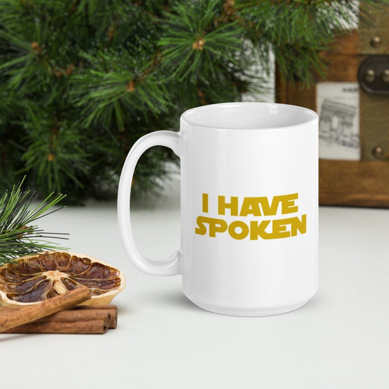 Puede incluir: Taza de cer&aacute;mica blanca con el texto "I HAVE SPOKEN" en letras doradas. La taza tiene un asa grande. El fondo incluye vegetaci&oacute;n y un cofre de madera.