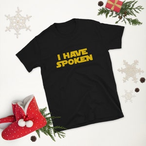 May include: Black short-sleeve t-shirt with the text "I HAVE SPOKEN" in yellow. The shirt is displayed on a white surface with holiday decorations, including a red gift box, pine needles, and a snowflake.
