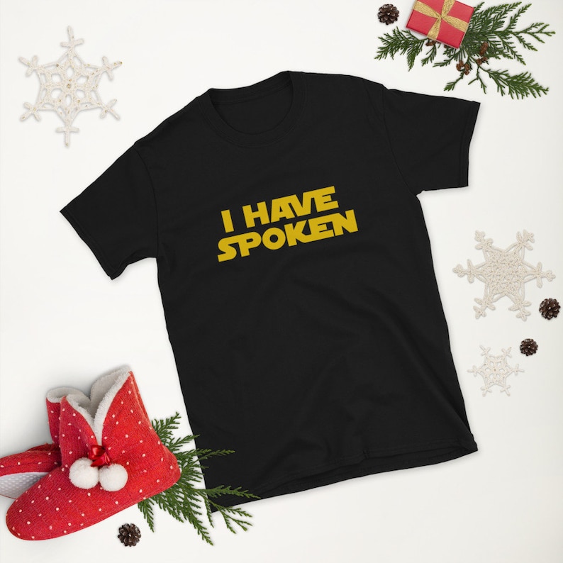 May include: Black short-sleeve t-shirt with the text "I HAVE SPOKEN" in yellow. The shirt is displayed on a white surface with holiday decorations, including a red gift box, pine needles, and a snowflake.
