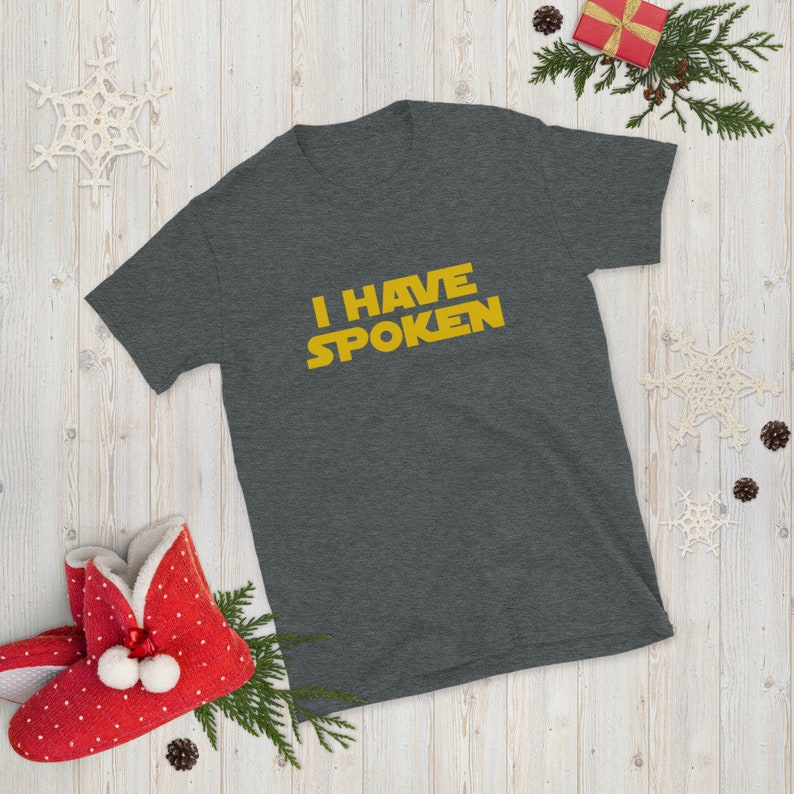 May include: Dark gray short-sleeve t-shirt with the text "I HAVE SPOKEN" in yellow. The shirt is displayed on a wooden surface with holiday decorations, including a red gift box and snowflake ornaments. A red slipper is also visible.